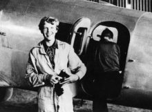 American aviator/pilot Amelia Earhart (1897-1937) standing by her Lockheed Electra dressed in overalls, with Fred Noonan getting into the plane in the background.